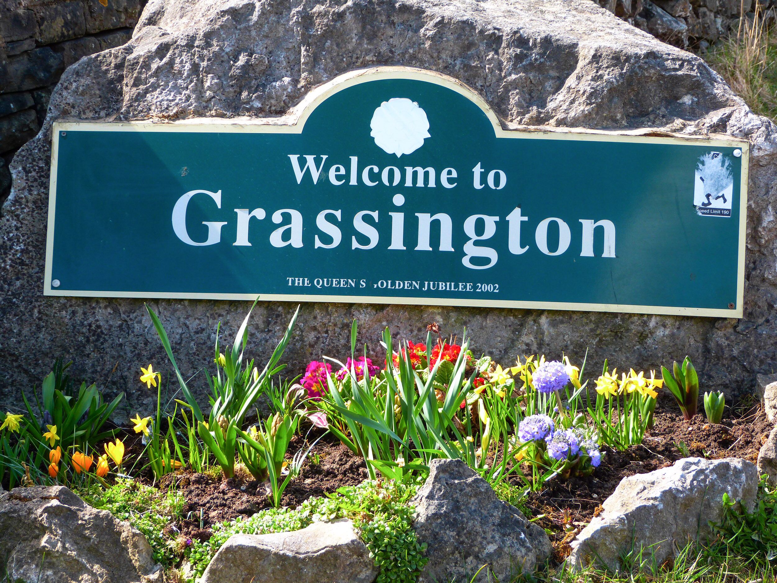 A welcome sign for Grassington with a speed limit sign and flowers in the foreground.