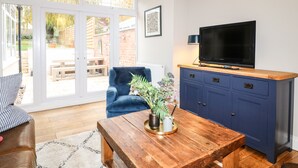 Uma sala de estar com mesa de centro de madeira, uma poltrona azul, televisão sobre um armário de madeira e uma porta de vidro deslizante que dá para um pátio externo.