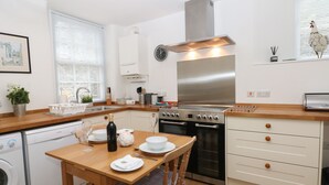 A compact kitchen with a built-in oven, a stainless steel range hood, and a wooden dining table with chairs. - 2B Cavendish Villas (Buxton)