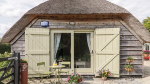 A rustic thatched-roof cottage with a wooden door and shuttered windows, a small table with chairs, and potted flowers on the porch. - The Barn at Rapps Cottage (Ilminster)