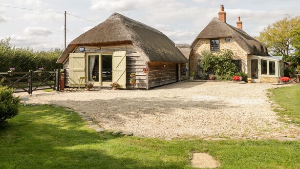 A traditional stone cottage with a thatched roof, a gravel driveway, and a small garden with flowers. - The Barn at Rapps Cottage (Ilminster)