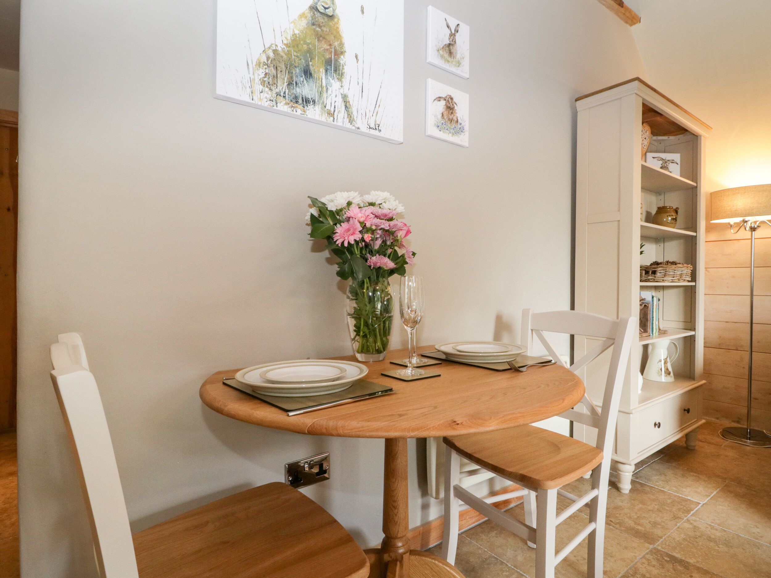 A dining area with a round wooden table, white chairs, a vase of flowers, and a white cabinet with shelves.