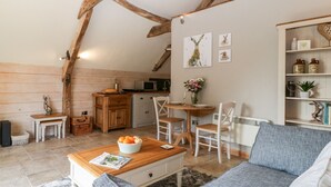 A cozy loft-style living space with exposed wooden beams, a dining area with a wooden table and chairs, a kitchen area, and a bookshelf. - The Barn at Rapps Cottage (Ilminster)