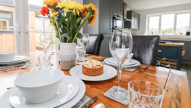 Una mesa de comedor dispuesta con platos y cuencos blancos, cubiertos y un pastel, con un florero en el centro.