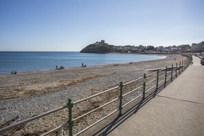 A pebble beach with a castle on a hill, a promenade, and buildings in the background. - Llawenfan (Criccieth)