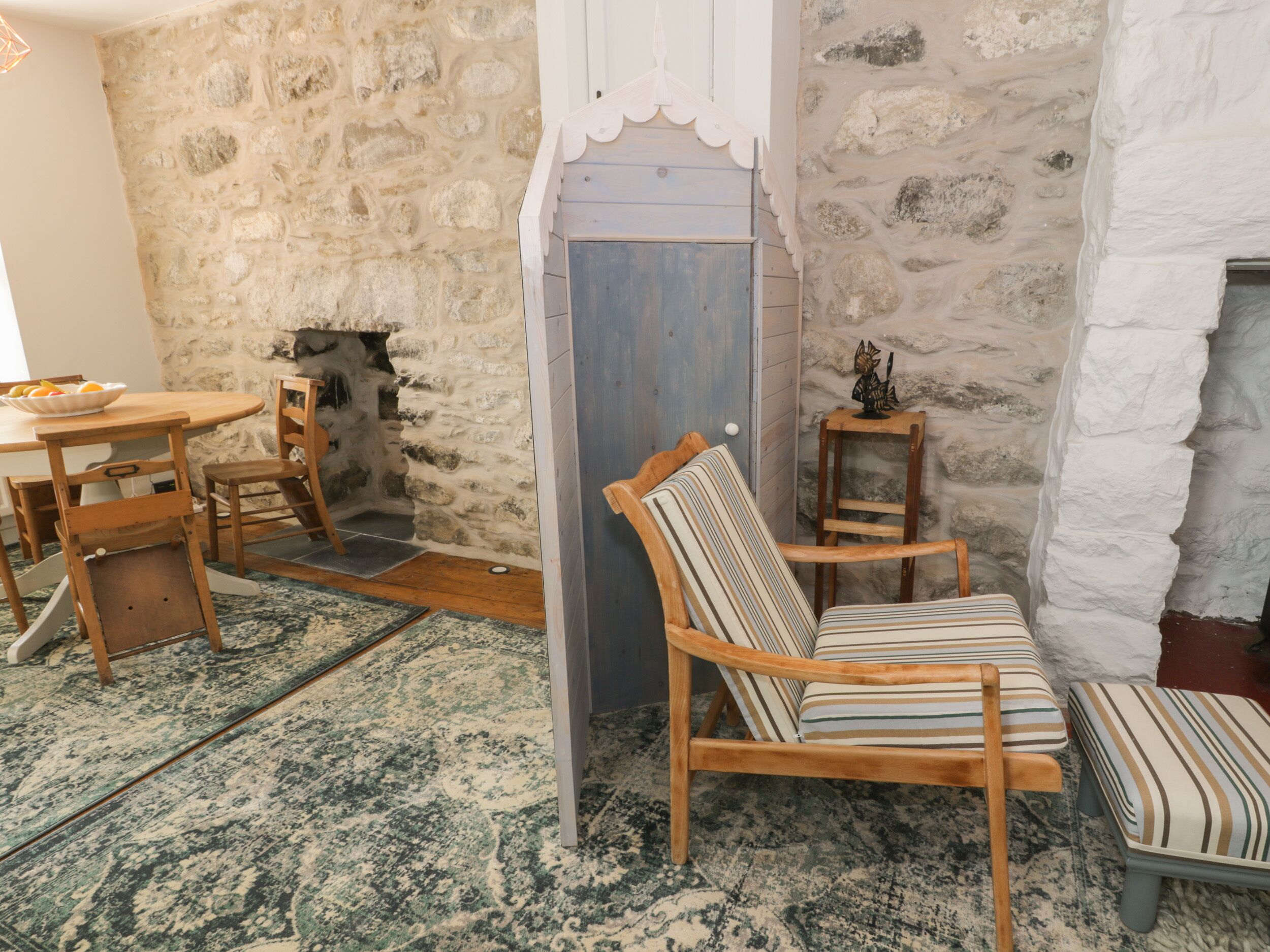 Um quarto acolhedor com parede de pedra, uma mesa de madeira com cadeiras, uma poltrona listrada e uma pequena mesinha com um objeto decorativo.
