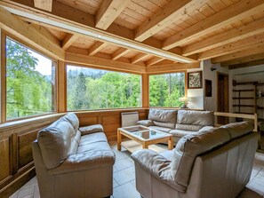 Ein Zimmer mit Holzwänden, einer Couch und einem Couchtisch, großen Fenstern mit Blick auf einen Wald und einem Bücherregal.