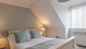 A bedroom with a sloped ceiling, a bed with white bedding and blue pillows, a bedside table with a lamp, and a window with blinds. - 4 Fishermans Wharf (Whitstable)