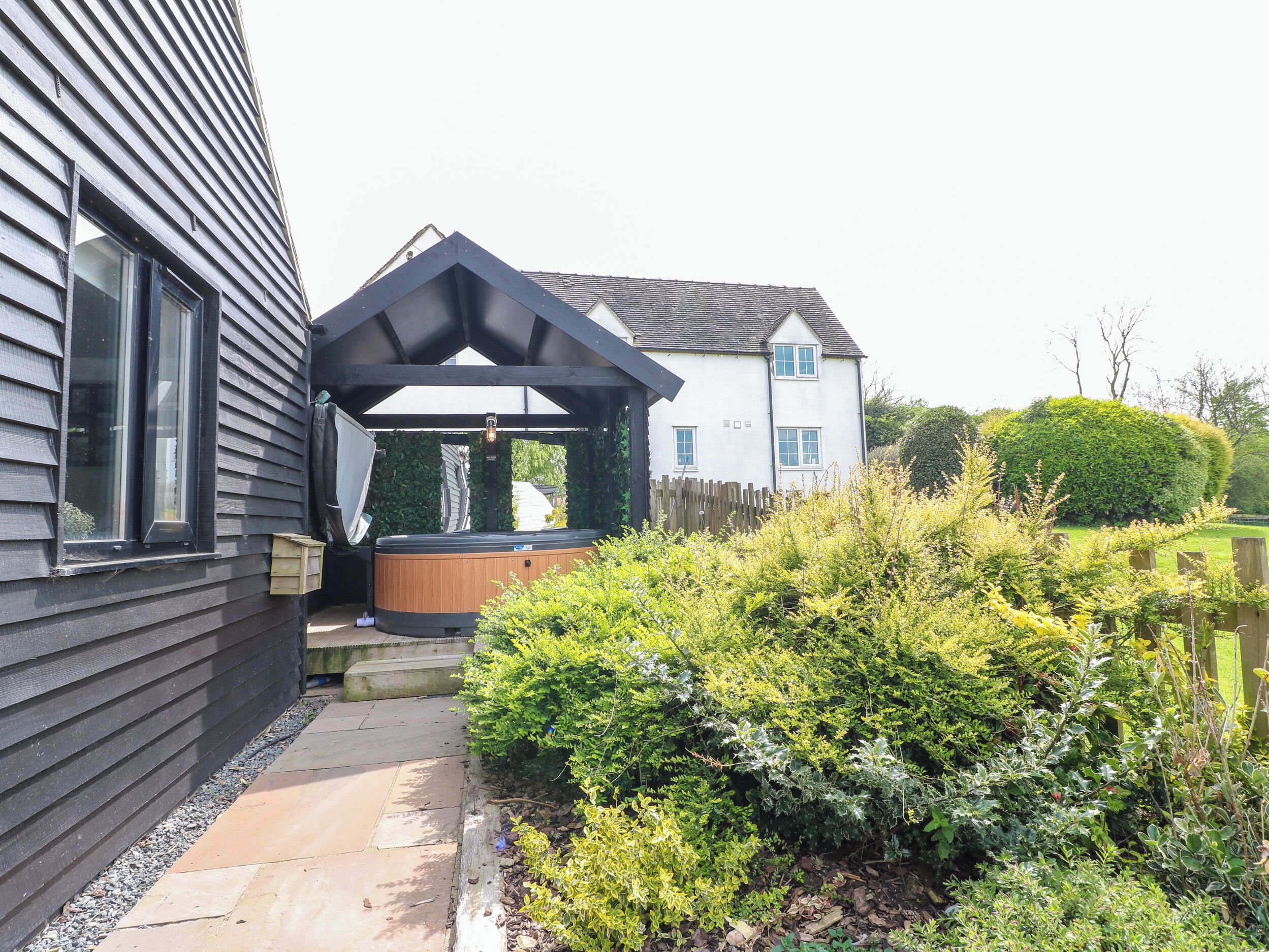 A hot tub under a wooden pergola adjacent to a house with a sloped roof and a fenced garden.