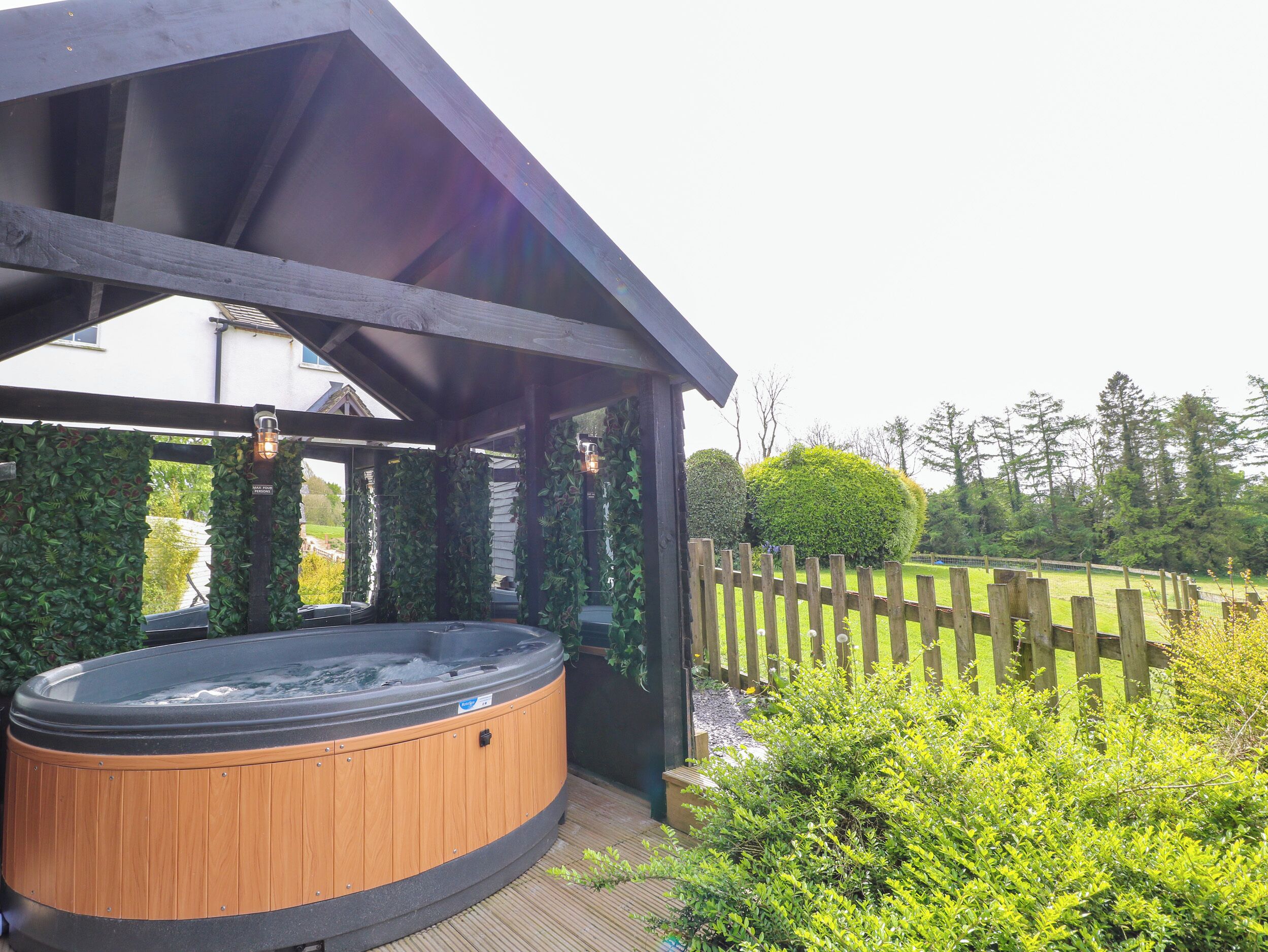 A hot tub under a wooden pergola with a wooden fence and greenery in the background.