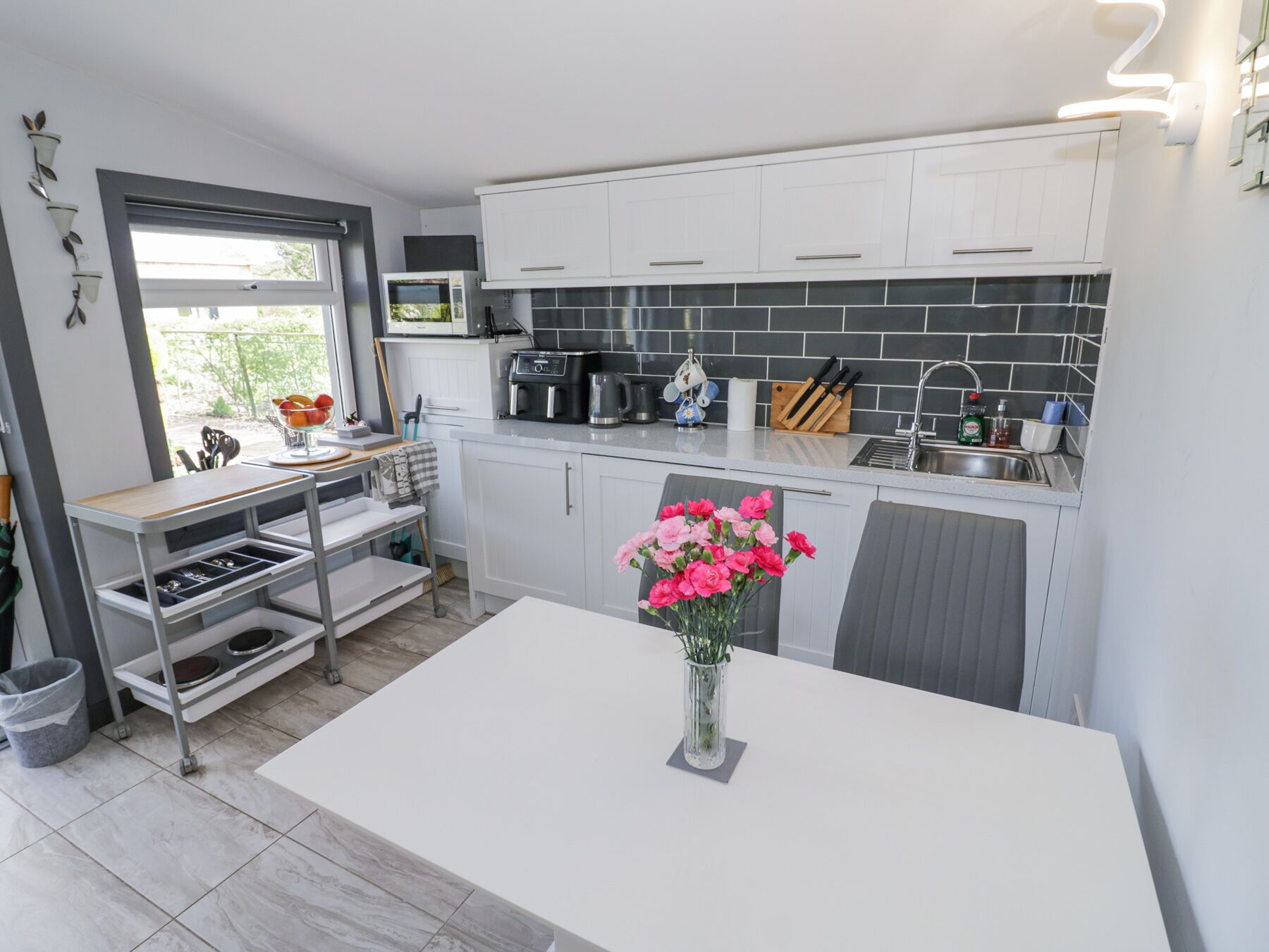 A compact kitchen with white cabinets, a black tiled backsplash, a coffee maker, and a vase of pink flowers on the counter.
