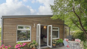 A wooden cabin with a patio, a dining area, and a garden with potted plants and a statue. - The Garden House (Stratford-upon-Avon)