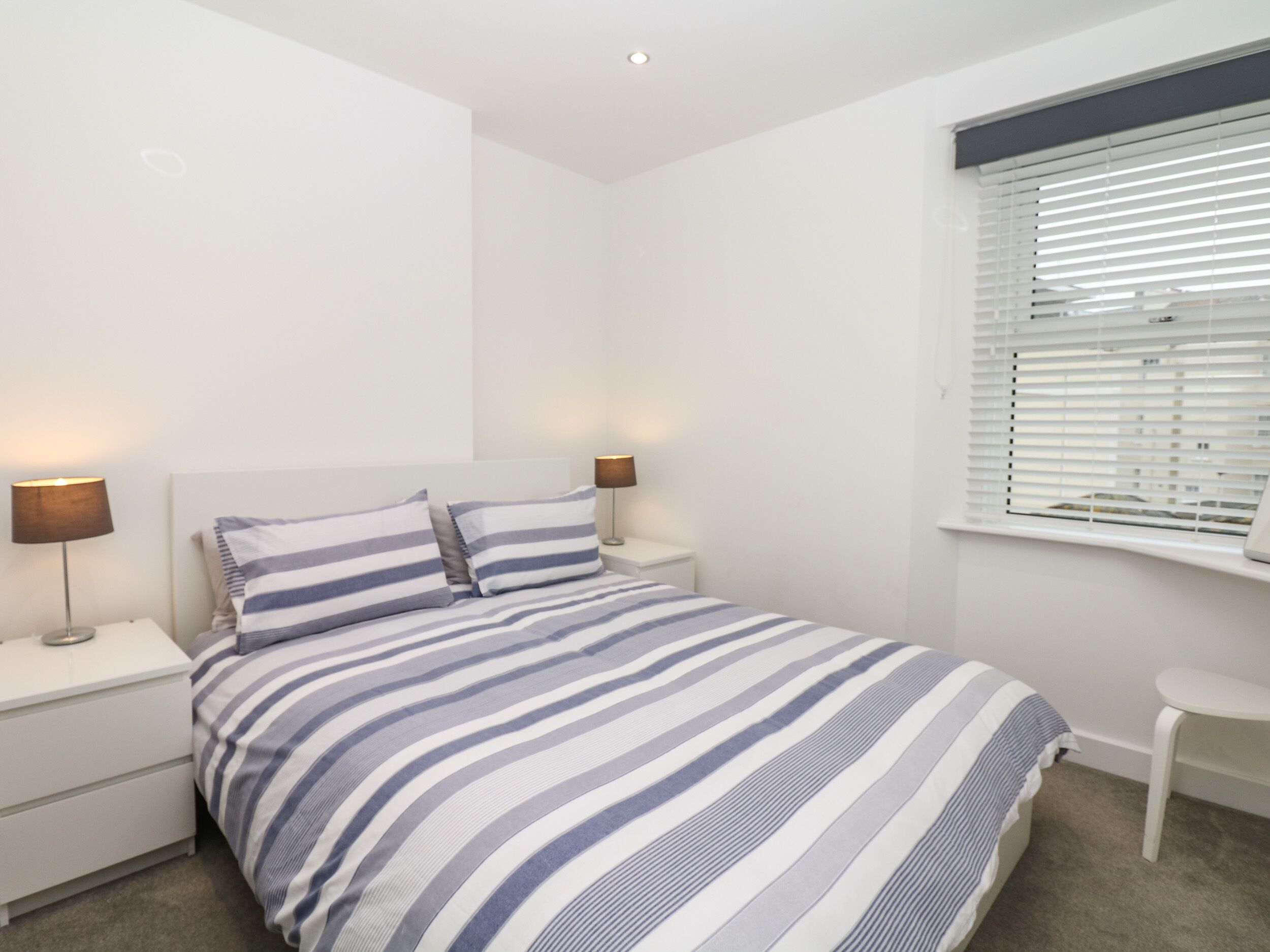 A bedroom with a bed, two bedside tables, two lamps, a window with blinds, and a small white chair.