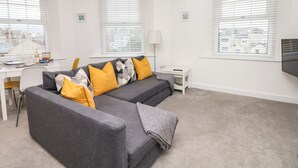A modern living room with a grey sofa, yellow and grey cushions, a small white side table, and a TV mounted on the wall. - Beach Rose (Teignmouth)