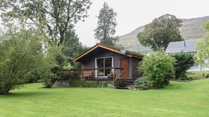 Une cabane en rondins avec un porche, entourée d’une cour verdoyante et d’arbres.