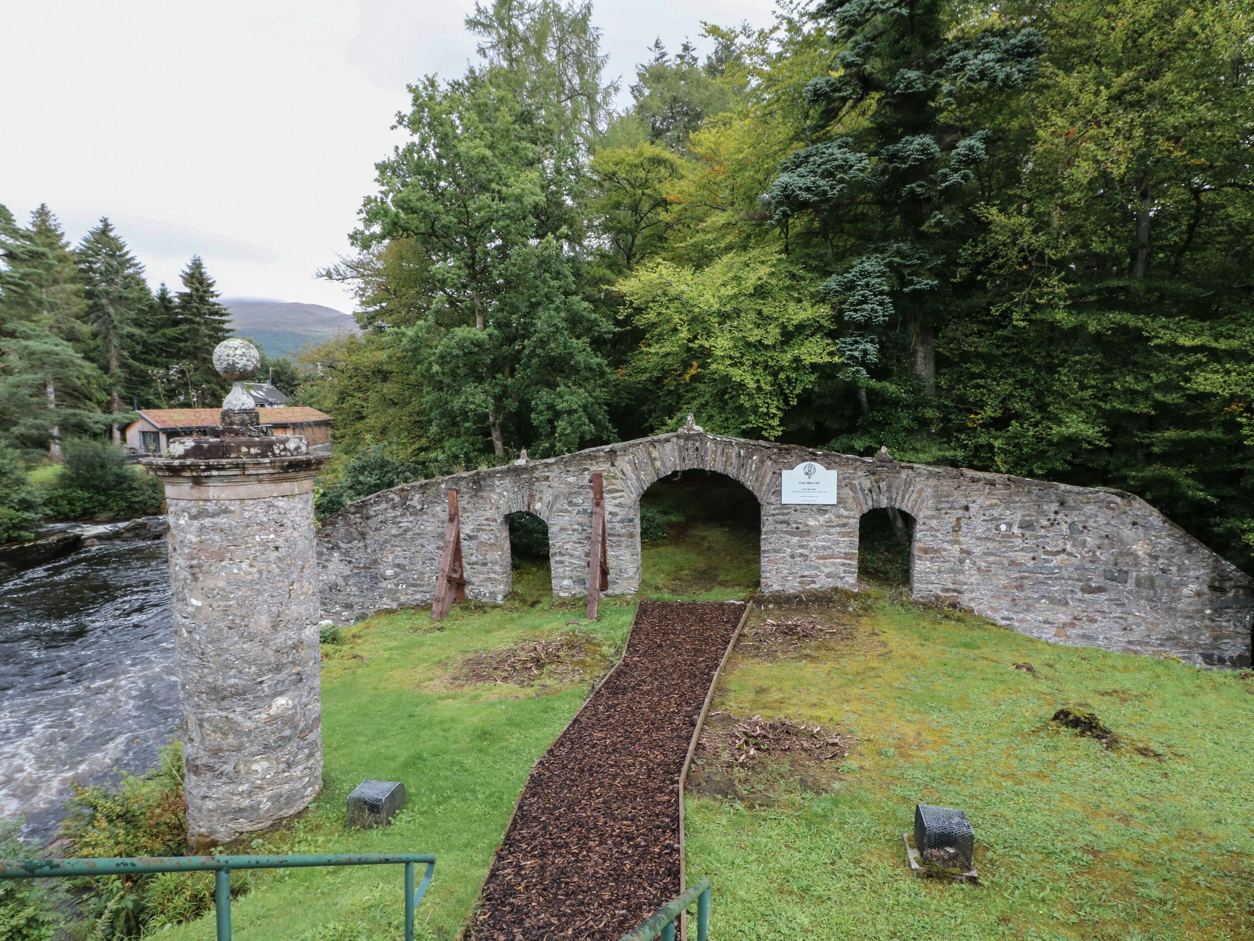 A stone bridge with three arches over a river, surrounded by greenery and a stone pillar with a spherical top.