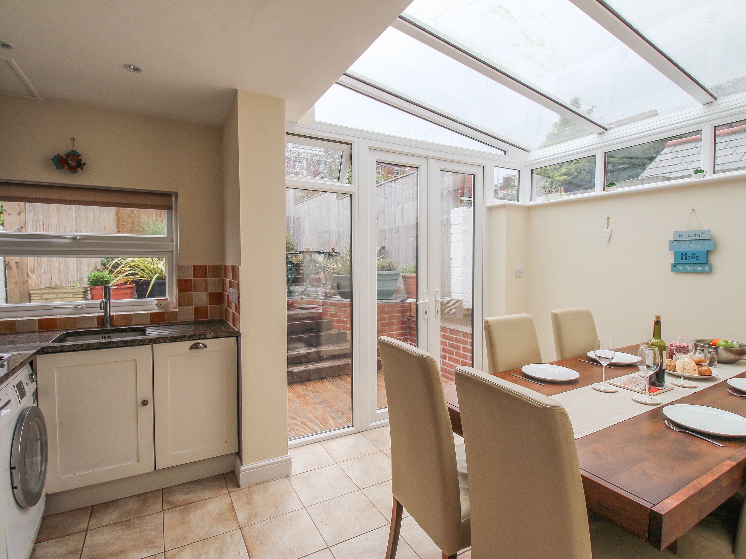 A kitchen with a built-in washing machine, a dining area with a table set for four, and a glass door leading to a patio with a view of a garden and a brick wall.