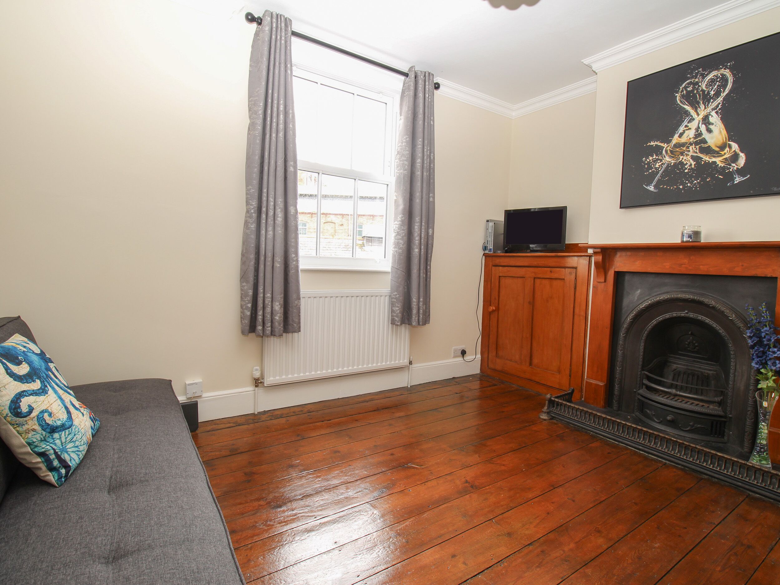 A living room with a wooden floor, a grey sofa, a fireplace, and a window with grey curtains.