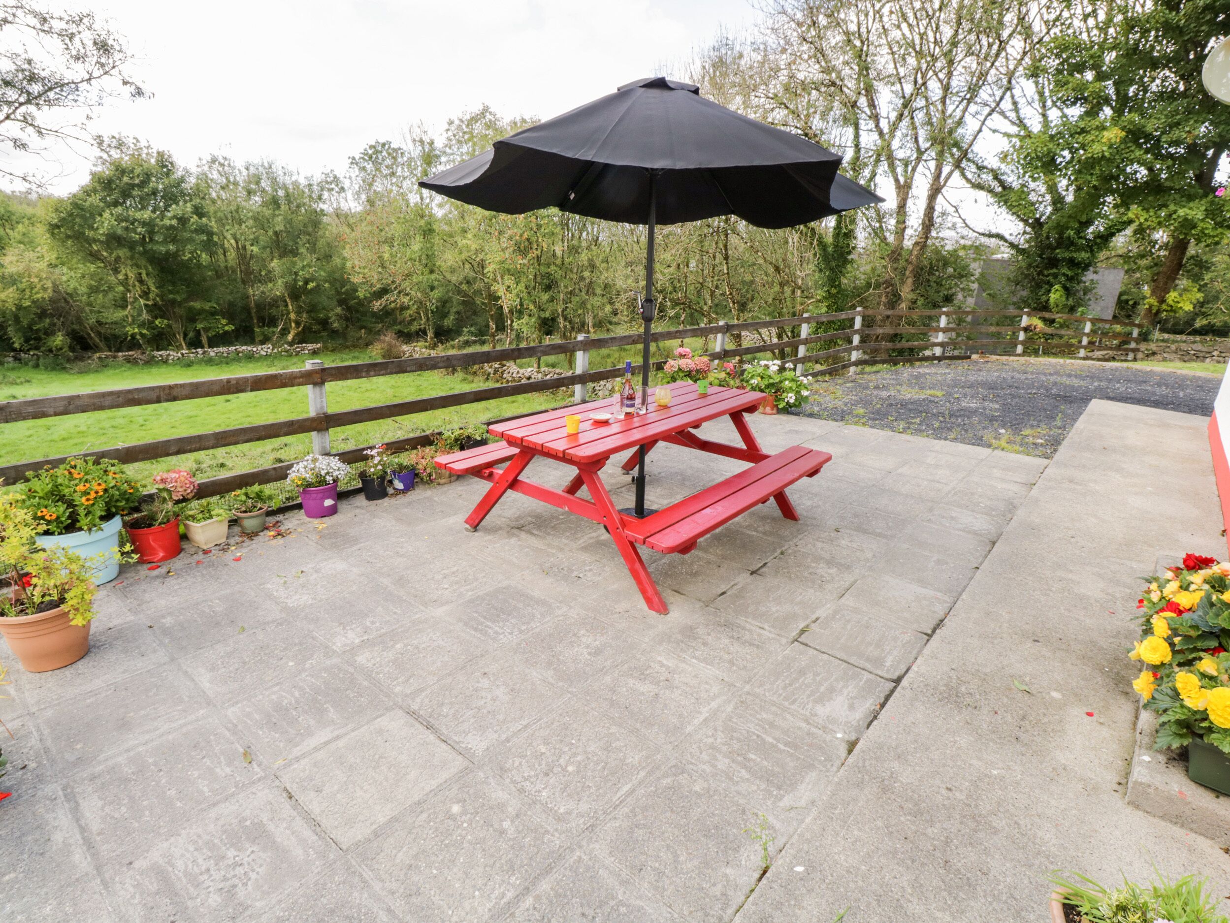 A red picnic table with a black umbrella, surrounded by potted plants and a wooden fence.