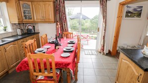A dining area with a table set for six, wooden chairs, a red tablecloth, and a bottle of wine. There is a window with curtains, a view of the outdoors, and a wooden cabinet with glass doors. - Lough Mask Road Fishing Cottage (Neale)
