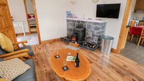 A cozy living room with a fireplace, a television mounted on the wall, and a dining area in the background. - Lough Mask Road Fishing Cottage (Neale)