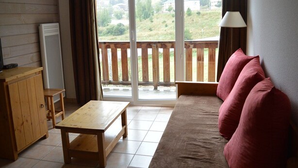 A cozy room with a wooden bench, a sofa with red cushions, a lamp, and a view of a balcony with a wooden railing. - T3 Apartment at the foot of the slopes with covered parking (Les Angles)