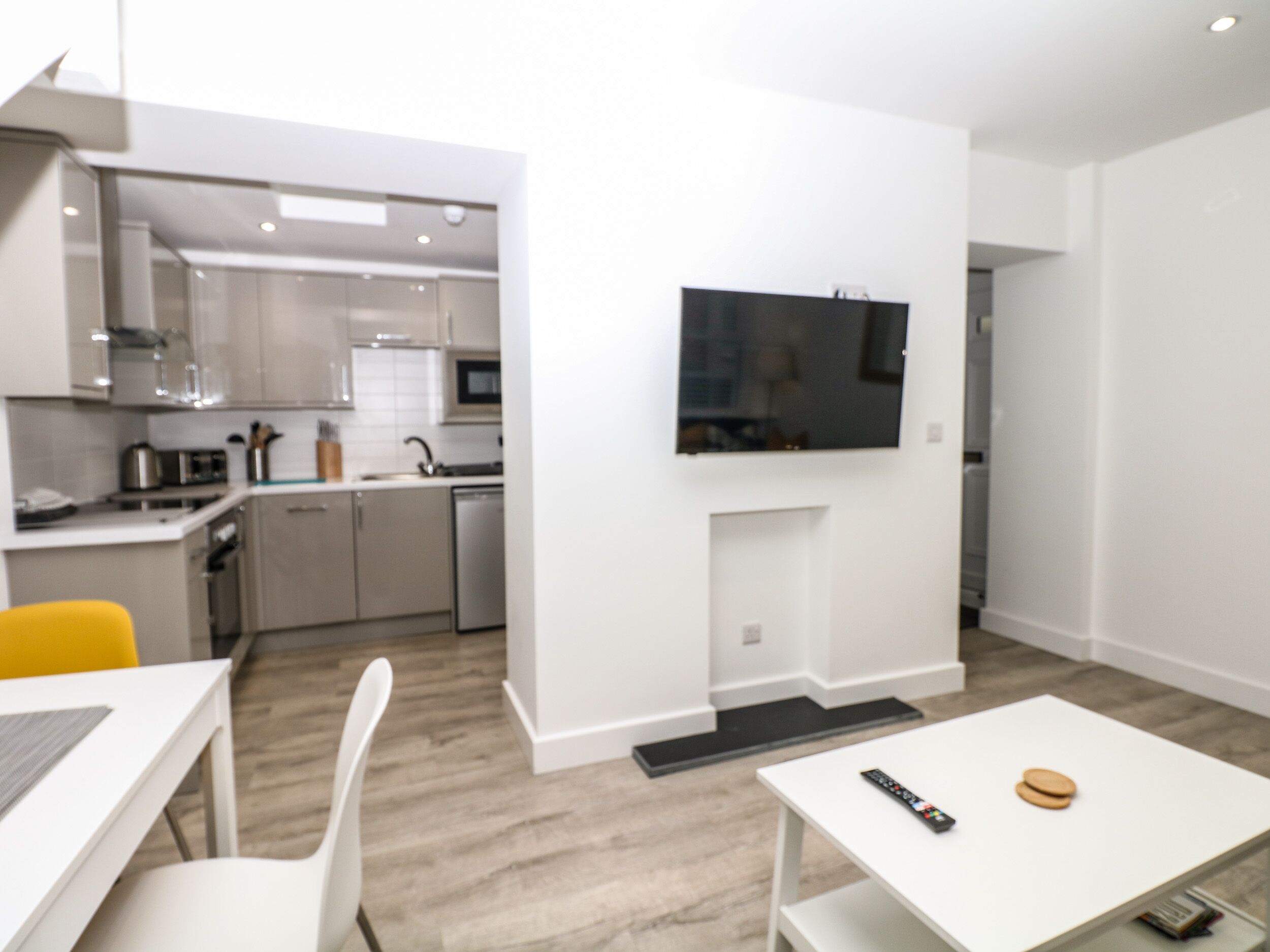 Una cocina moderna con armarios blancos, un microondas y un lavamanos. Hay una zona de comedor con una mesa y sillas blancas. Un televisor de pantalla plana está montado en la pared sobre una chimenea.