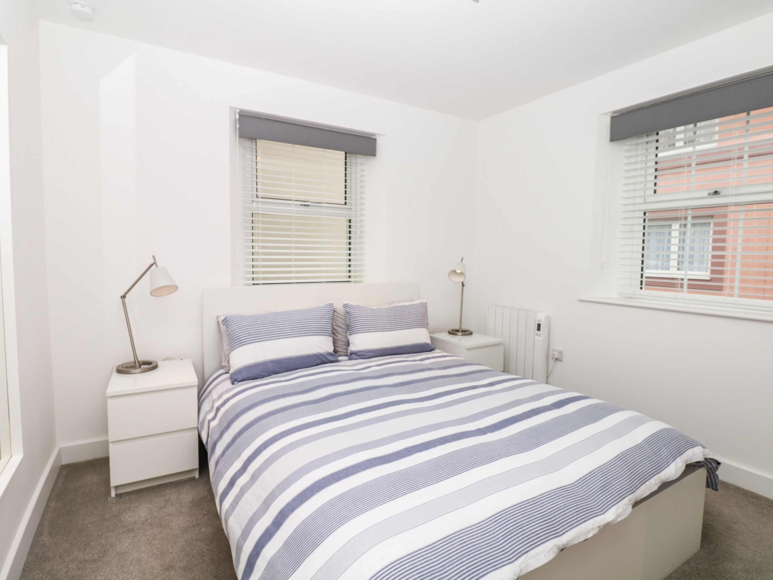 Une chambre à coucher avec un lit, deux tables de chevet, une fenêtre avec des stores et un radiateur.