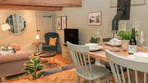 Un salon chaleureux avec un canapé, une table basse ronde et un téléviseur. On y trouve également un coin repas avec une table dressée pour deux personnes, une chaise et une bouteille de vin.