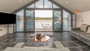 A spacious living room with a large glass door leading to a balcony overlooking a body of water. - Boathouse on the Lake (Windermere)