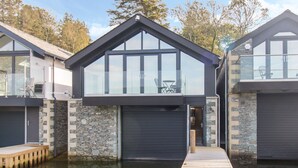 A modern house with a large glass window, a stone facade, and a wooden dock extending into a body of water. - Boathouse on the Lake (Windermere)