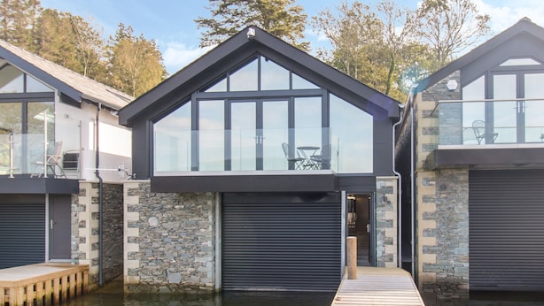 A modern house with a large glass window, a stone facade, and a wooden dock extending into a body of water. - Boathouse on the Lake (Windermere)