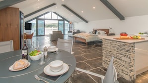 A spacious loft-style bedroom with a large bed, a dining area with a table set for two, and a kitchen with a stone island. - Boathouse on the Lake (Windermere)