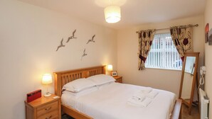 A neatly made bed with white linens, a wooden nightstand with a lamp, a bedside table with a red object, and a window with patterned curtains. - Cadliam (Whitby)