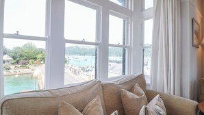 Een kamer met een groot raam met uitzicht op een jachthaven en een wateroppervlak, een beige bank met meerdere kussens, en een klein tafeltje.