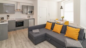 A modern kitchen with grey cabinets, a microwave, and a dishwasher. There is a grey sofa with yellow cushions in the living area, and a window with blinds. - Samphire (Teignmouth)