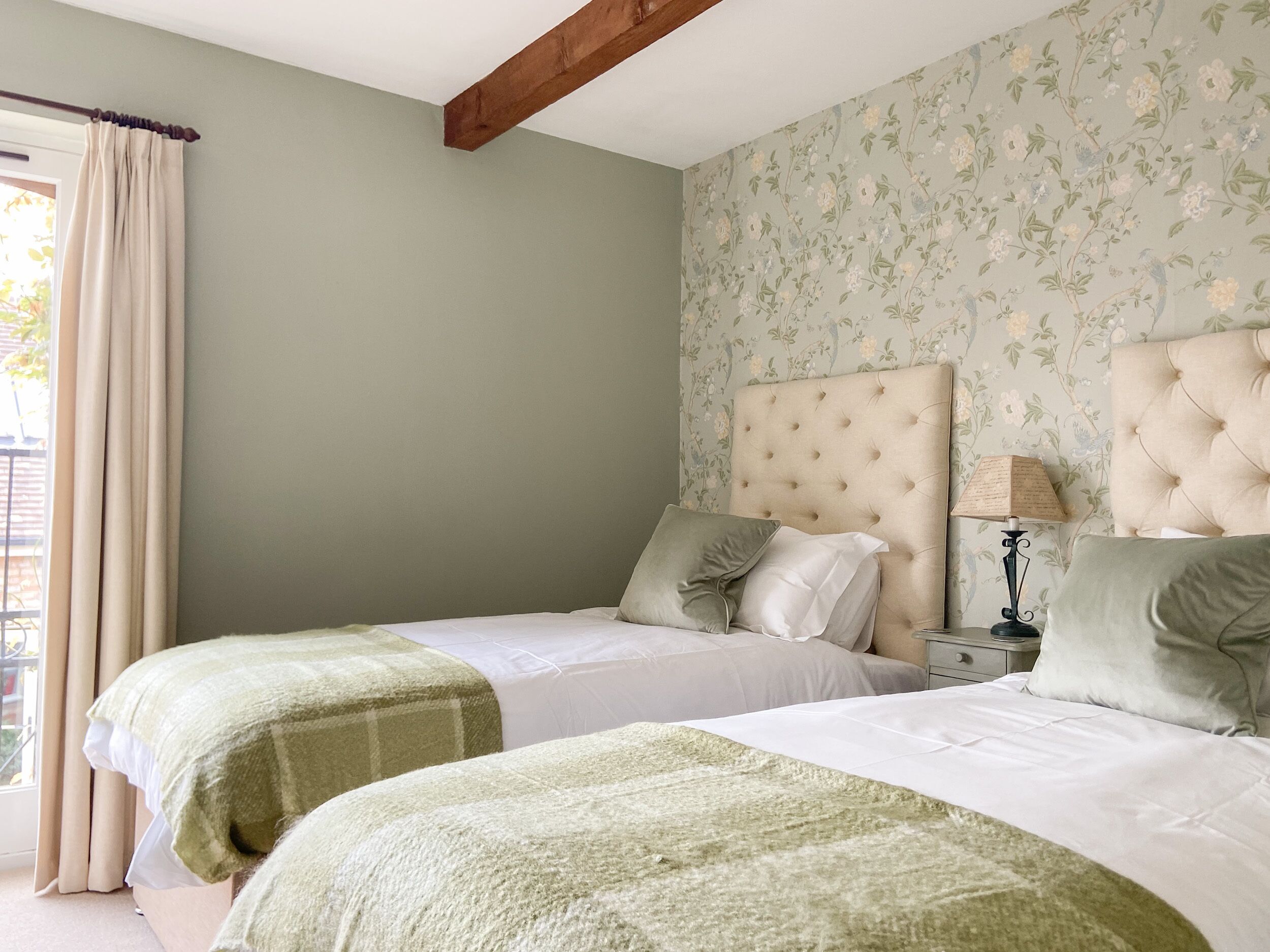 Un dormitorio con dos camas, papel tapiz floral, un viguelo de madera y una ventana con cortinas.
