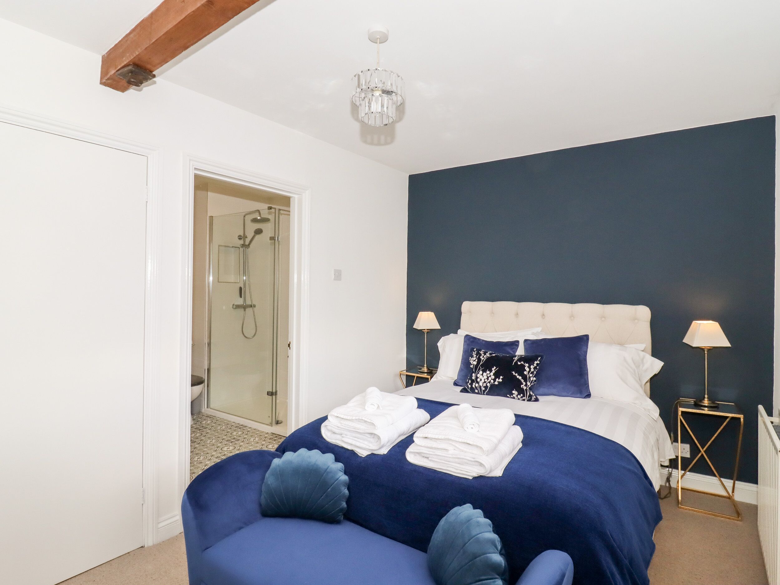 A bedroom with a bed, two bedside tables, a nightstand, a chandelier, and a glass shower door.