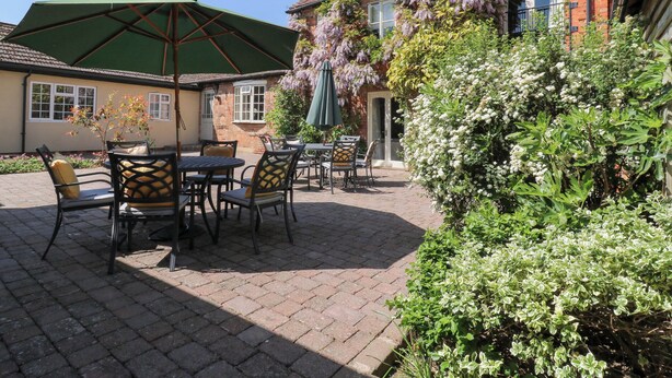 Eine Terrasse im Freien mit Tischen und Stühlen, einem grünen Sonnenschirm und einem Backsteinhaus mit Ziegeldach.