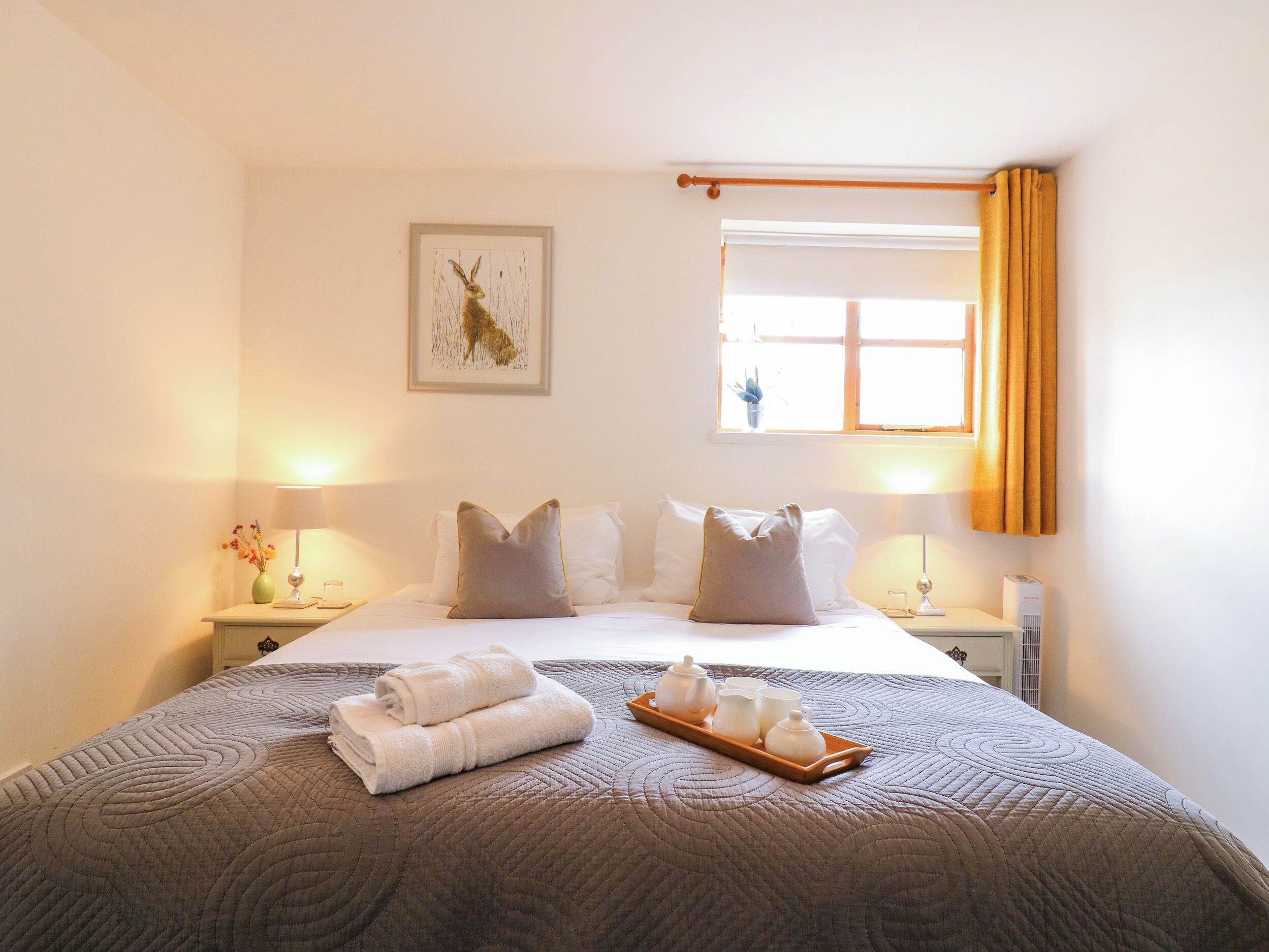 A hotel room with a bed, two bedside tables, a window with curtains, a wall-mounted artwork, and a tray with tea sets.