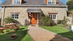 A stone house with a wooden front door, two windows, and a gravel pathway leading to a wooden table and chairs. - The Linhay (Axminster)