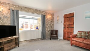 A living room with a stone wall, a television on a wooden stand, a brown leather chair, a brown sofa, and a wooden door. - The Linhay (Axminster)