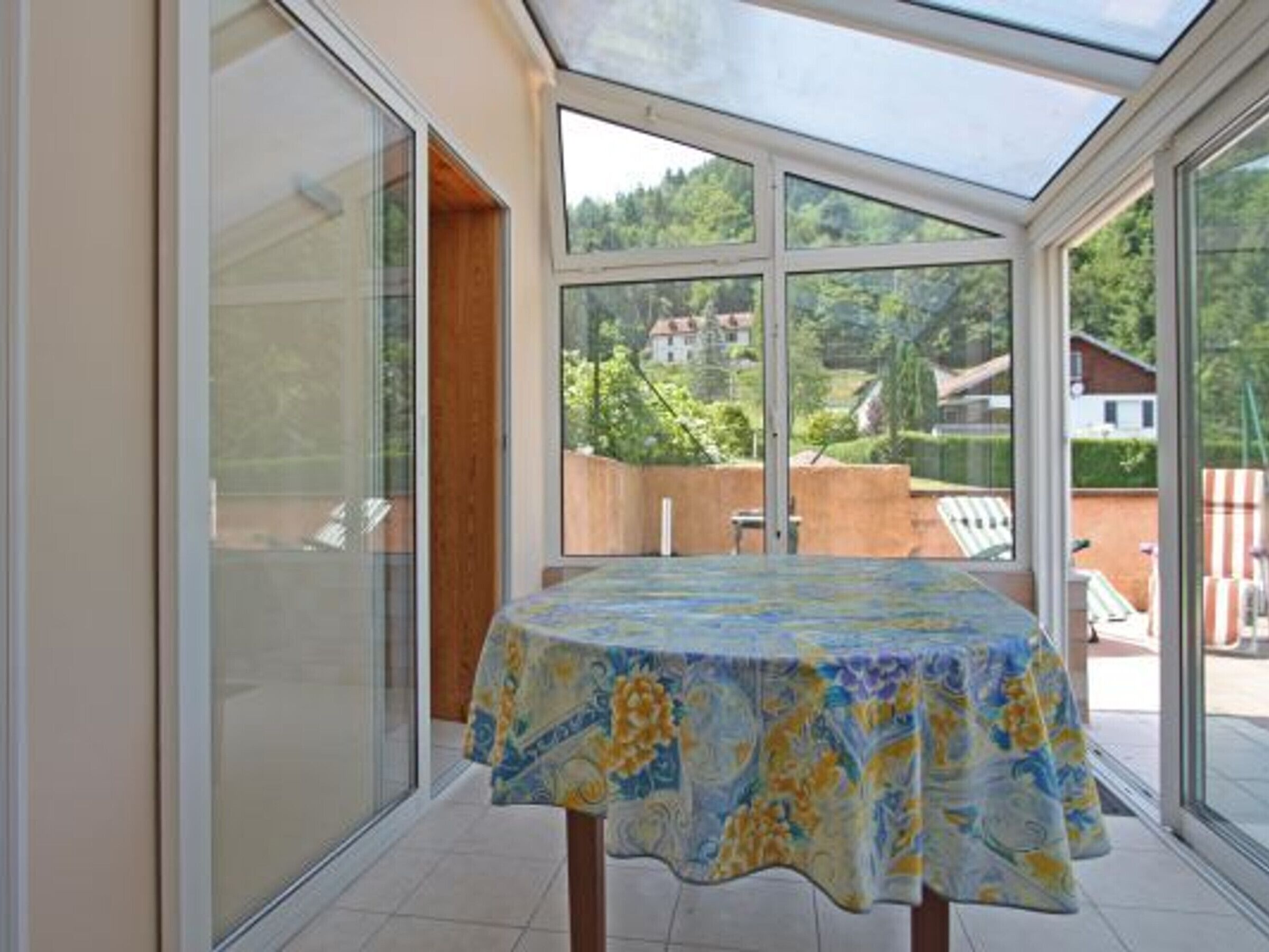 Een kamer met een tafel bedekt met een bloemendessin, grote glasramen en uitzicht op een woongebied.