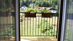 A balcony with a metal railing, a wooden planter box with flowers, and a view of a green landscape with houses in the background. - Cozy Village Retreat with Private Pond, Terrace, and Family Amenities in Le Ménil (Le Menil)