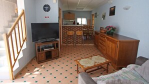 A living room with a TV, wooden furniture, a staircase, and a kitchen in the background. - Cozy Village Retreat with Private Pond, Terrace, and Family Amenities in Le Ménil (Le Menil)