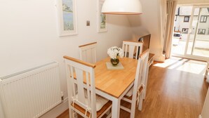 A dining area with a wooden table and white chairs, a hanging pendant light, and framed artwork on the walls. - Ketch Cottage (Holyhead)