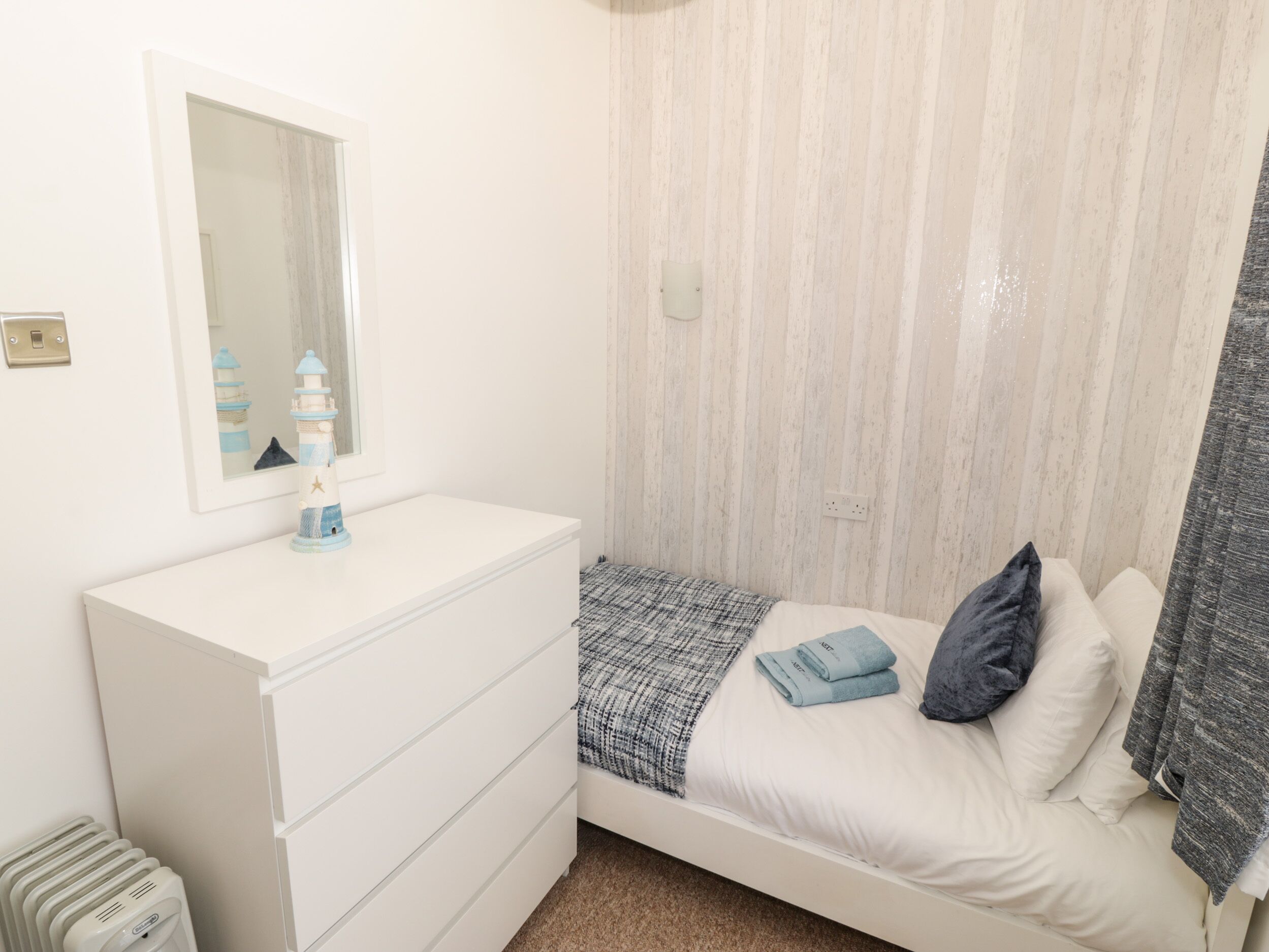 Een slaapkamer met een witte dressoir, een bed met een blauwe deken en kussens, een spiegel boven het dressoir, en een gestreepte muur.