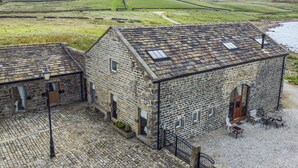 A stone building with a tiled roof, a small outdoor seating area, and a view of rolling hills and a body of water. - Snave Barn (Skipton)