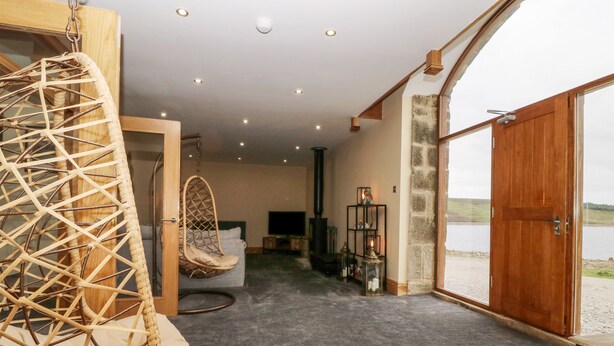 A cozy room with a hanging chair, a sofa, a fireplace, and a large window overlooking a body of water. - Snave Barn (Skipton)