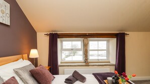 A hotel room with a bed, pillows, a bedside lamp, a window with curtains, a radiator, and a tray with a mug, flowers, and a pastry. - Mirk Cottage (Hebden Bridge)